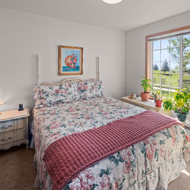 A cozy bedroom with a floral-patterned bedspread and matching pillows on a double bed. A pink knitted throw blanket is draped across the foot of the bed. There is a nightstand with a lamp and a small device on the left side of the bed. A framed painting of flowers hangs on the wall above the bed. To the right, a large window shows a green outdoor landscape with trees and patio furniture. Several potted plants are placed on a white dresser beneath the window.