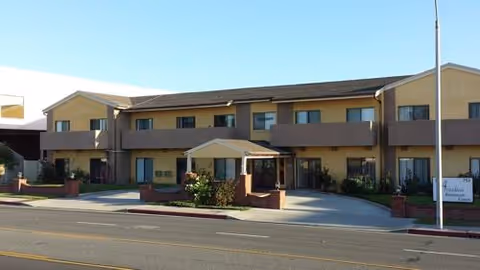 Two-story beige retirement center with a covered entrance, balconies, and landscaping along the front.