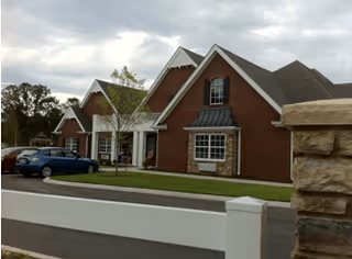 Exterior view of a brick senior living facility building with multiple peaked roofs, windows, and a small parking area with cars. There is a white fence in the foreground and some trees in the background under a cloudy sky.