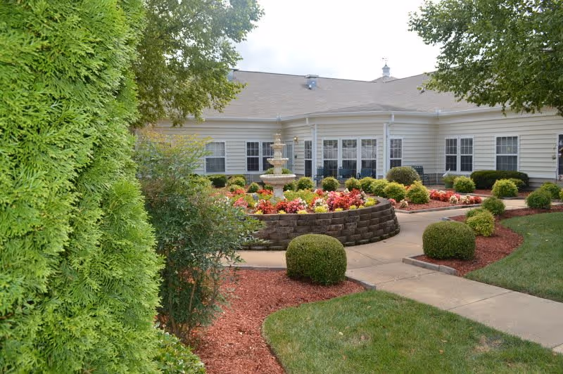 Outdoor garden area at Morning Pointe of Columbia featuring a circular flower bed with a multi-tiered fountain in the center, surrounded by neatly trimmed bushes, colorful flowers, and a paved walkway. The building with multiple windows is visible in the background under a partly cloudy sky.