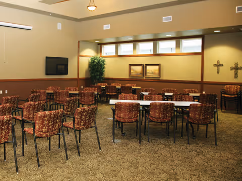 A spacious room with multiple tables and chairs arranged in rows, patterned carpet flooring, beige walls with decorative crosses and framed pictures, a flat screen TV mounted on the wall, and a potted plant in the corner.