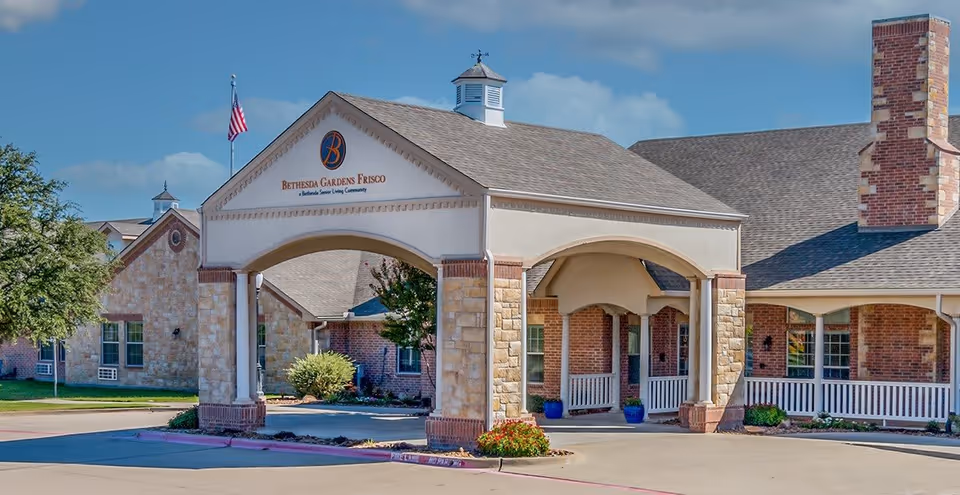 Front exterior view of Bethesda Gardens Frisco Assisted Living facility showing a covered entrance with stone and brick pillars, an American flag, and a clear blue sky.