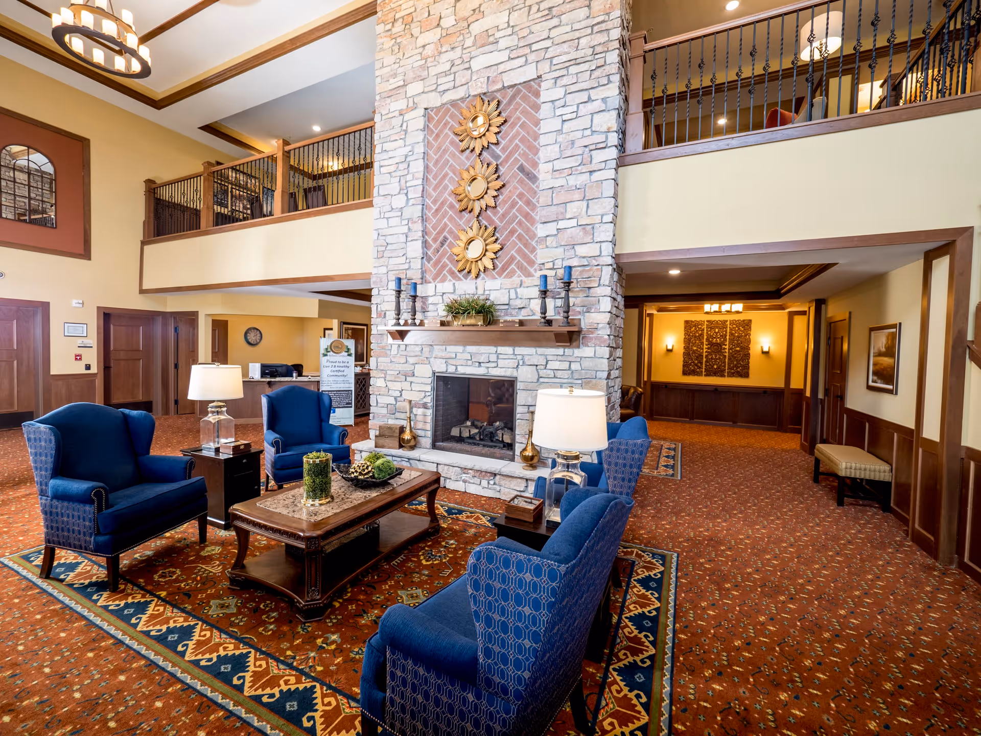 A spacious senior living facility common area with a large stone fireplace in the center. Surrounding the fireplace are four blue upholstered armchairs and a wooden coffee table with decorative items. The room features a patterned carpet, warm yellow walls, and a high ceiling with a chandelier. There is a balcony with wrought iron railings overlooking the area and a hallway leading to other rooms.