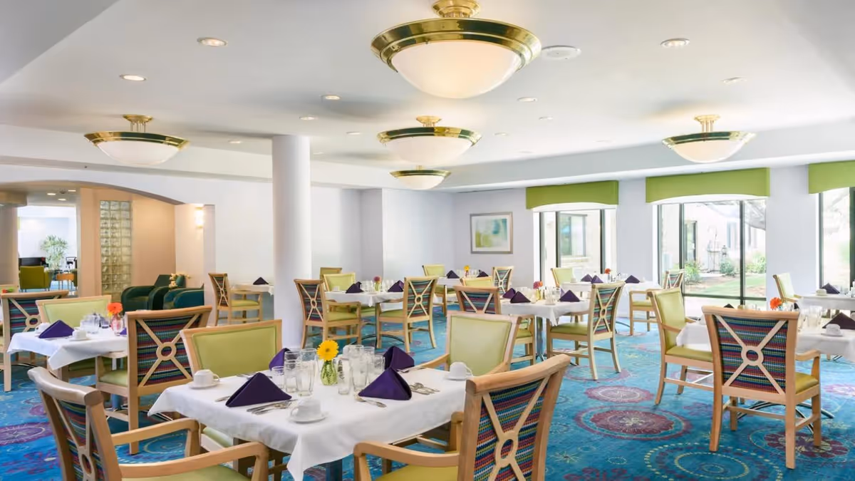 Bright dining room with multiple tables covered in white tablecloths, each set with purple napkins, cups, glasses, and small flower vases. The room has large windows with green valances letting in natural light, colorful patterned chairs, and blue carpet with circular designs. Ceiling lights with gold accents illuminate the space.