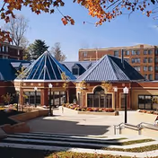 Brick senior living building front with glass pyramid roof structures, a paved courtyard with steps and planters, and autumn foliage overhead.