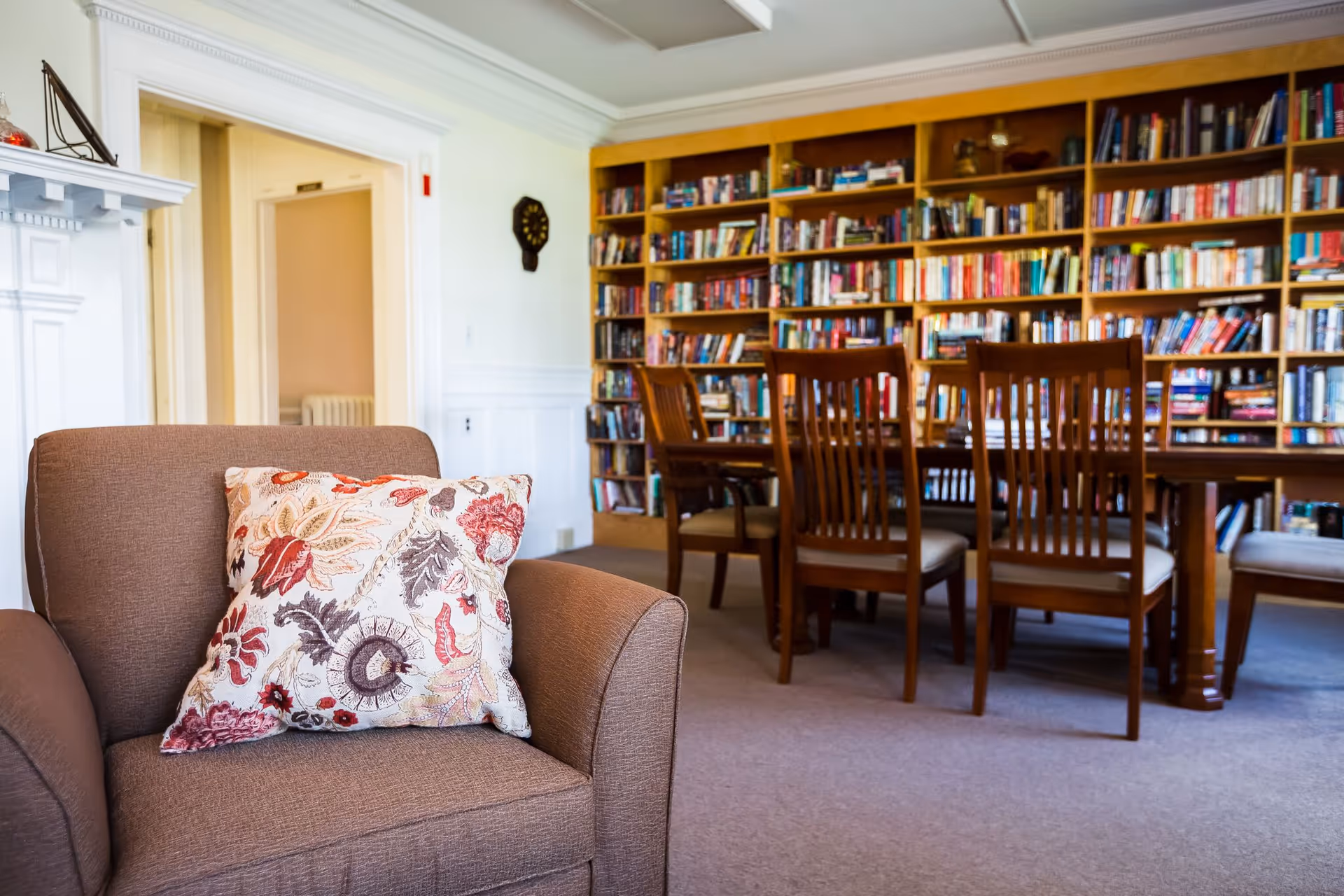 Cozy interior room with a brown armchair featuring a floral patterned cushion in the foreground. In the background, there is a large wooden bookshelf filled with books and a wooden table surrounded by chairs. The room has white walls with decorative molding and a wall clock.