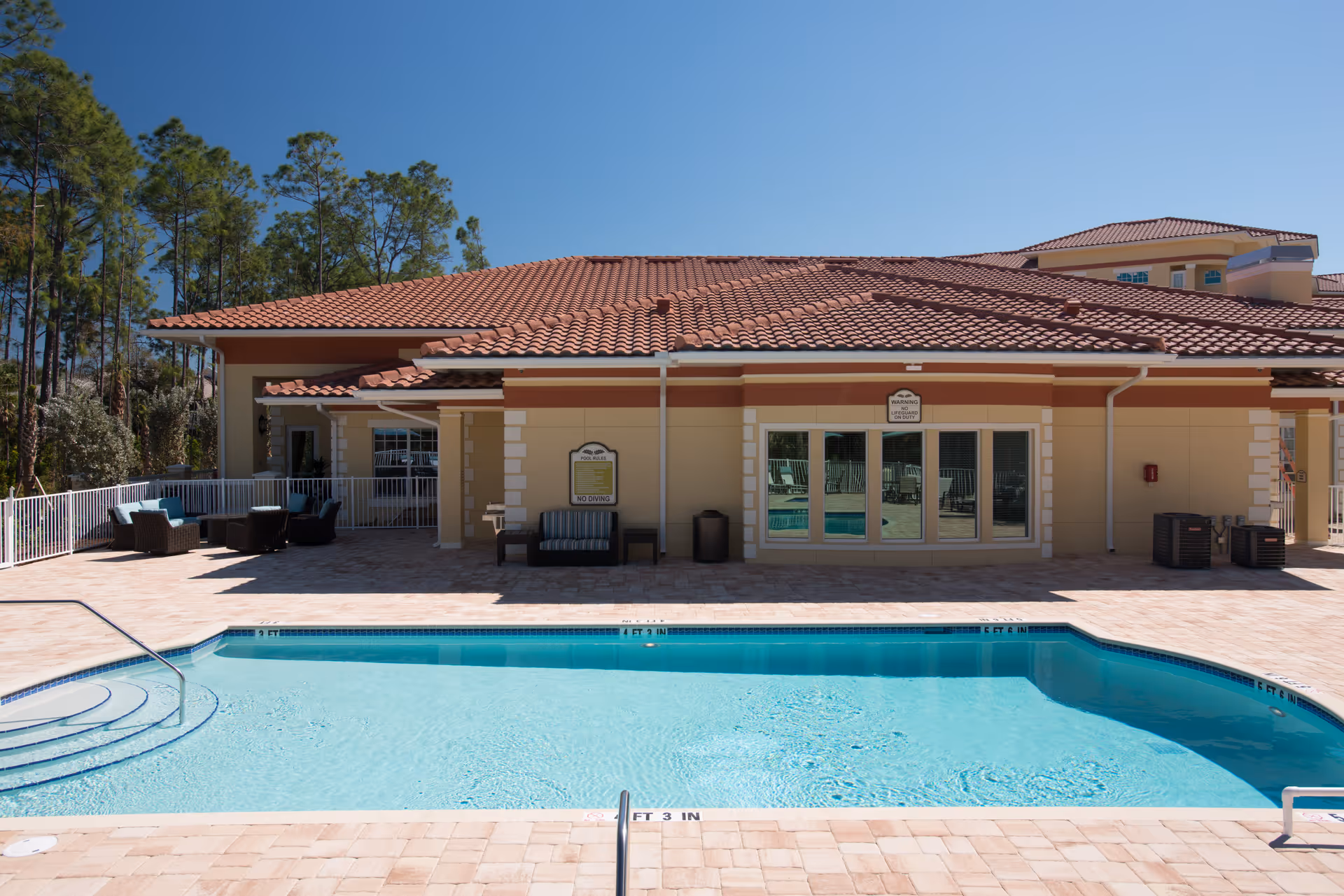 Outdoor swimming pool with clear blue water in front of a building with a red tiled roof. There are patio chairs and tables on the pool deck, surrounded by a white fence. Trees are visible in the background under a clear blue sky.