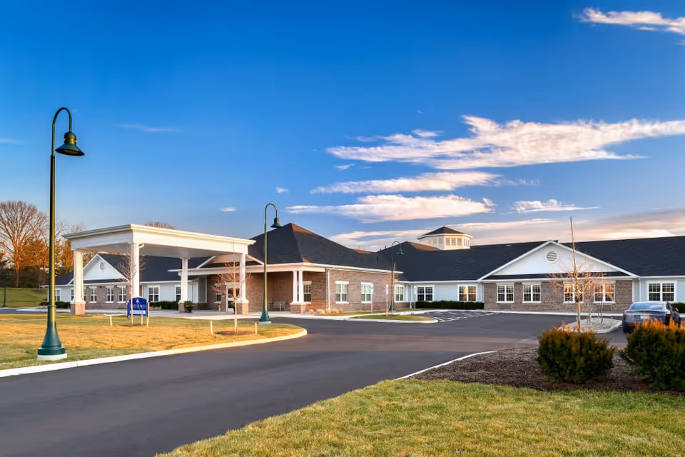 Front exterior of a single-story brick-and-white-siding senior living building with a covered porte-cochère, driveway, lamp posts, and landscaped lawn under a blue sky.