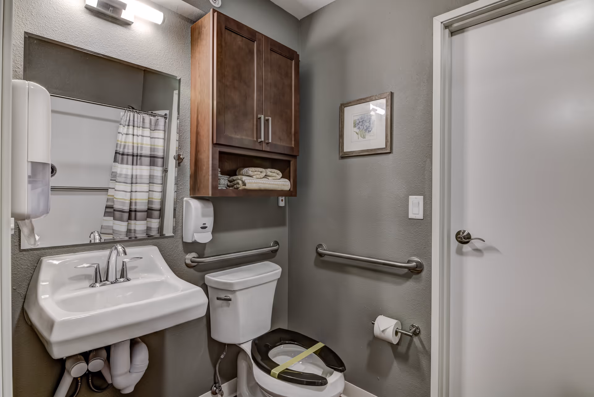 A small bathroom with a white sink and chrome faucet, a toilet with a black seat secured by yellow tape, a wooden cabinet mounted above the toilet with folded towels inside, a wall-mounted soap dispenser, grab bars on the walls, a framed floral picture, and a shower with a striped curtain visible in the mirror reflection.