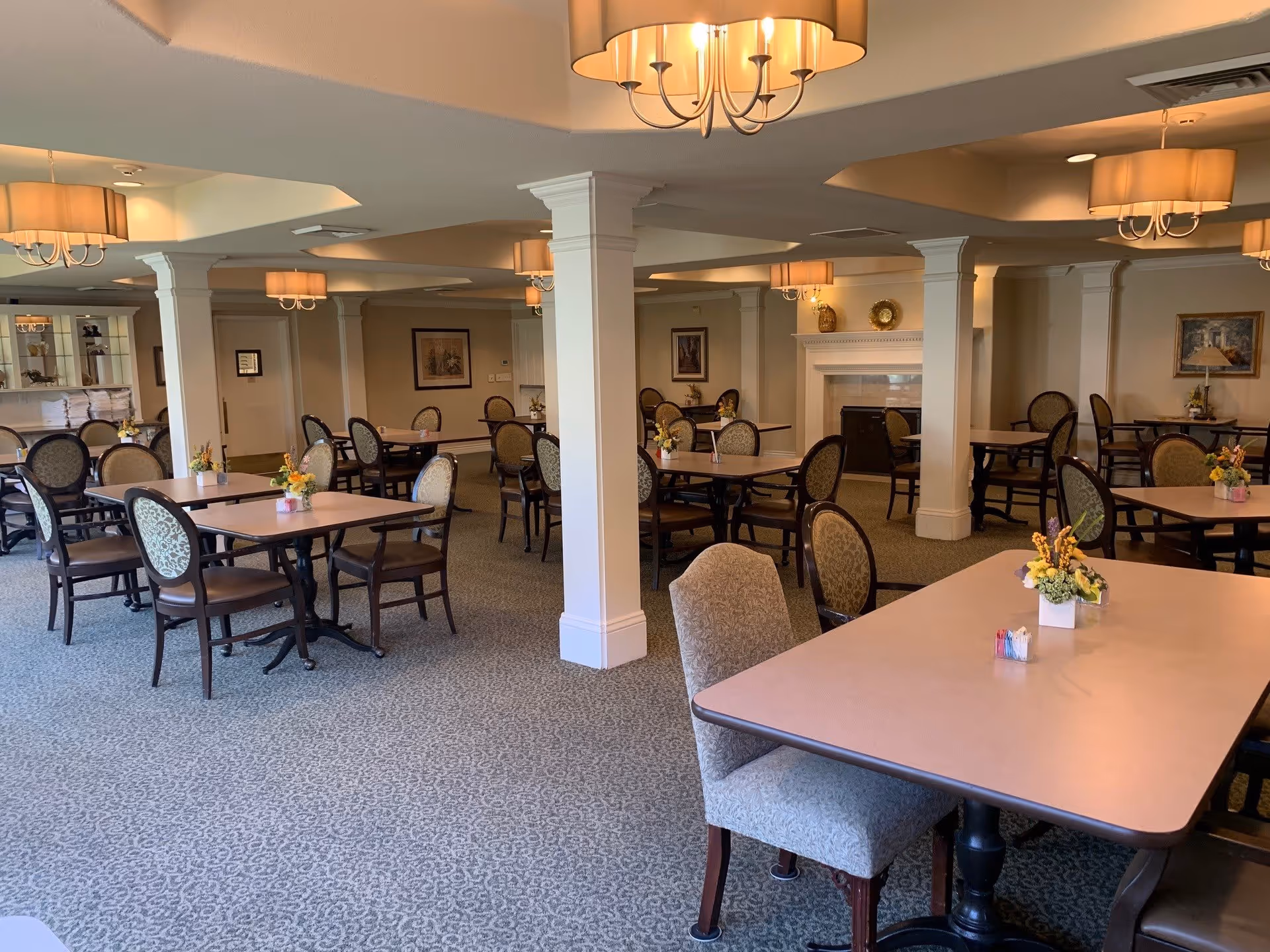 A spacious dining room with multiple tables and chairs arranged neatly. Each table has a small floral centerpiece. The room features beige walls, carpeted flooring, white columns, and several ceiling light fixtures with lampshades. There is a fireplace on one side of the room and framed artwork on the walls.