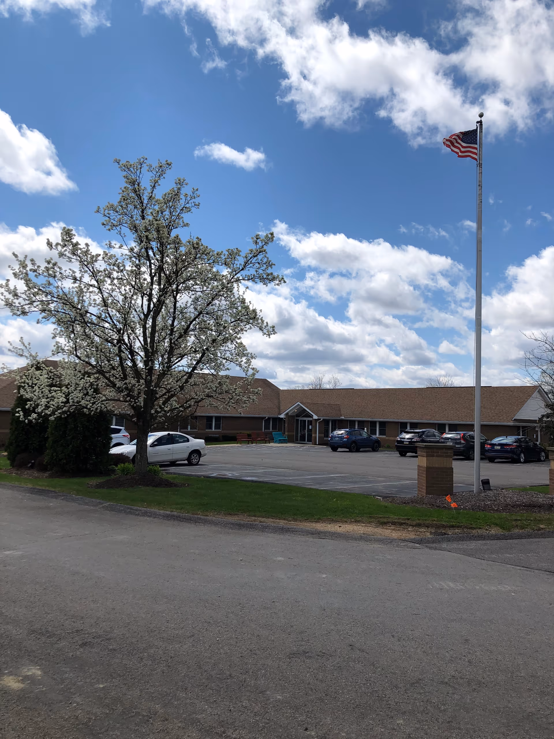 Exterior view of a single-story building with a brown roof and brick facade under a partly cloudy blue sky. There is a parking lot with several cars, a blooming tree with white flowers, and an American flag on a tall flagpole in the foreground.
