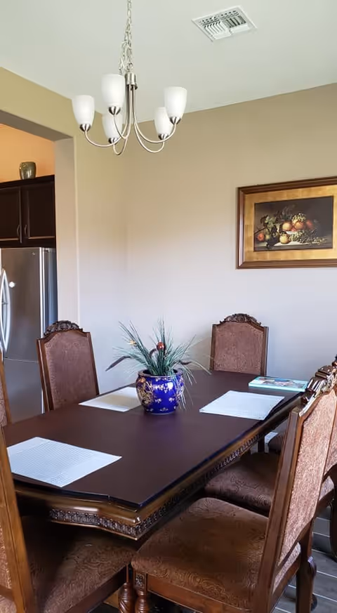 A dining room with a dark wooden table surrounded by six upholstered chairs. The table has white placemats and a blue decorative vase with artificial plants in the center. A framed painting of fruit hangs on the beige wall. A modern chandelier with five lights hangs from the ceiling. A stainless steel refrigerator is partially visible through an open doorway to the left.