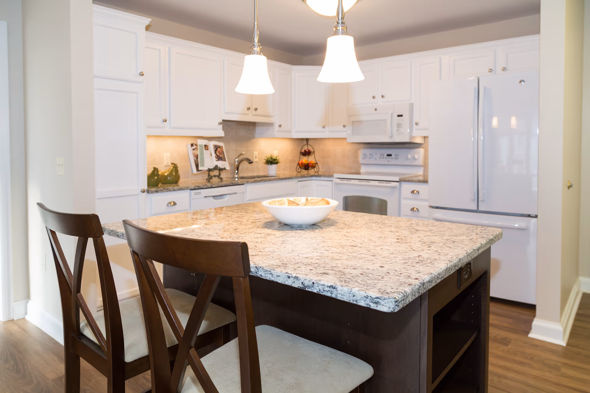 A modern kitchen with white cabinets, a white refrigerator, a white stove with a microwave above it, and a granite countertop island with two wooden chairs. Two pendant lights hang above the island, which has a white bowl with bread on it. The floor is wooden and the backsplash is tiled in a neutral color.
