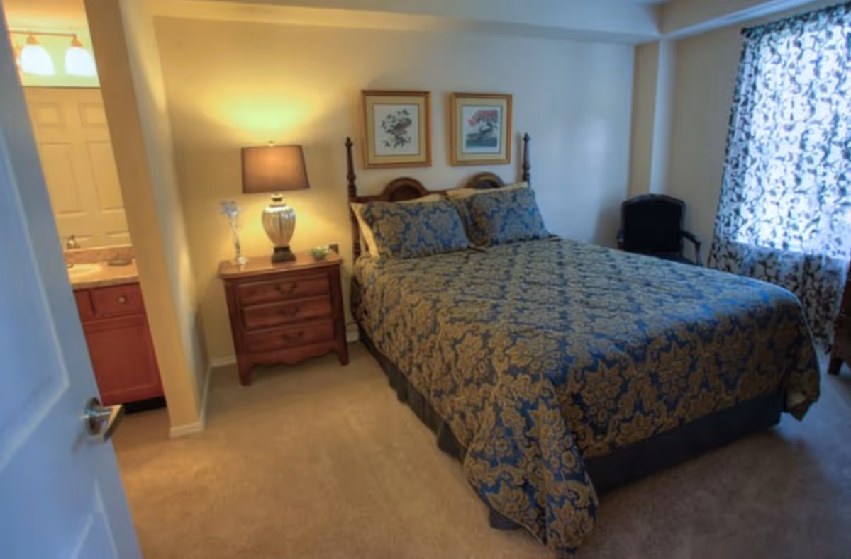 A bedroom with a large bed covered in a blue and gold patterned bedspread. There is a wooden nightstand with a lamp and decorative items next to the bed. Two framed pictures hang on the wall above the bed. A window with patterned curtains lets in natural light, and a black chair is positioned near the window. The door to an adjoining bathroom is open, showing a sink and mirror.