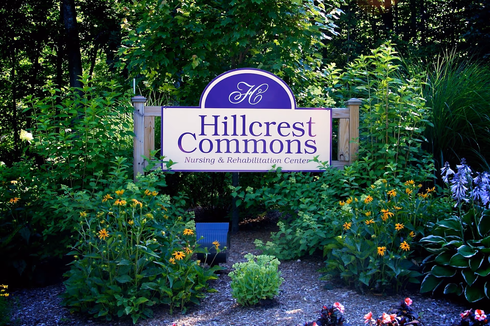 A wooden sign for Hillcrest Commons Nursing & Rehabilitation Center surrounded by lush green plants and colorful flowers in a garden setting.