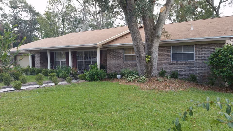 Single-story brick building with a brown roof surrounded by green grass, shrubs, and trees. The building has several windows and a covered porch area with chairs.
