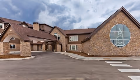 Exterior view of a senior living facility building with a brick facade, a covered entrance supported by stone pillars, multiple windows, and a large circular window displaying a statue inside. The sky is cloudy and there is a paved driveway and crosswalk in front.