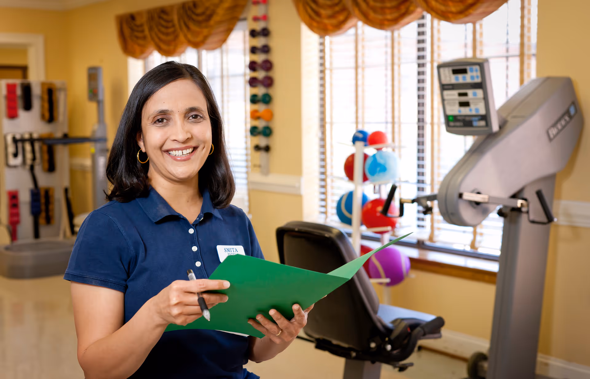 A smiling woman wearing a navy blue polo shirt and holding a green folder and pen stands in a well-lit room with exercise equipment and colorful therapy balls in the background.