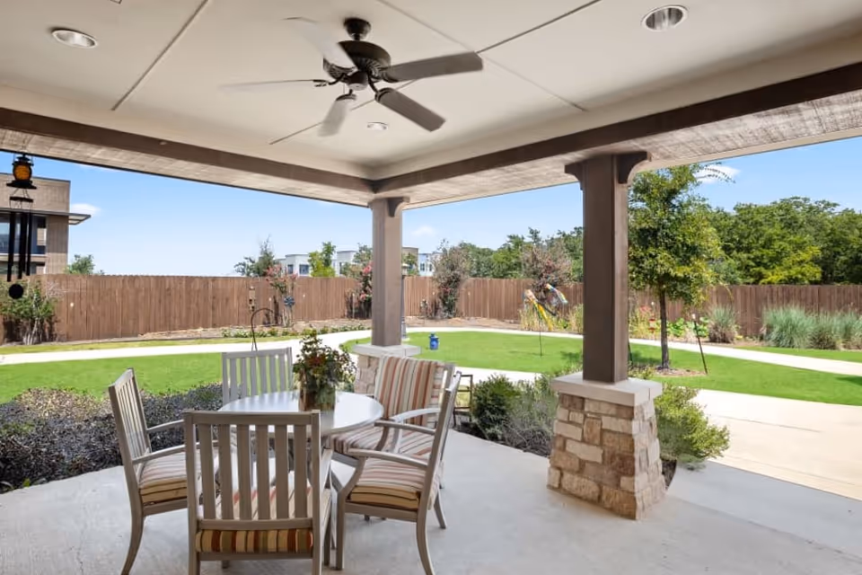 Covered patio with a round table and four striped chairs overlooking a fenced lawn and garden path.