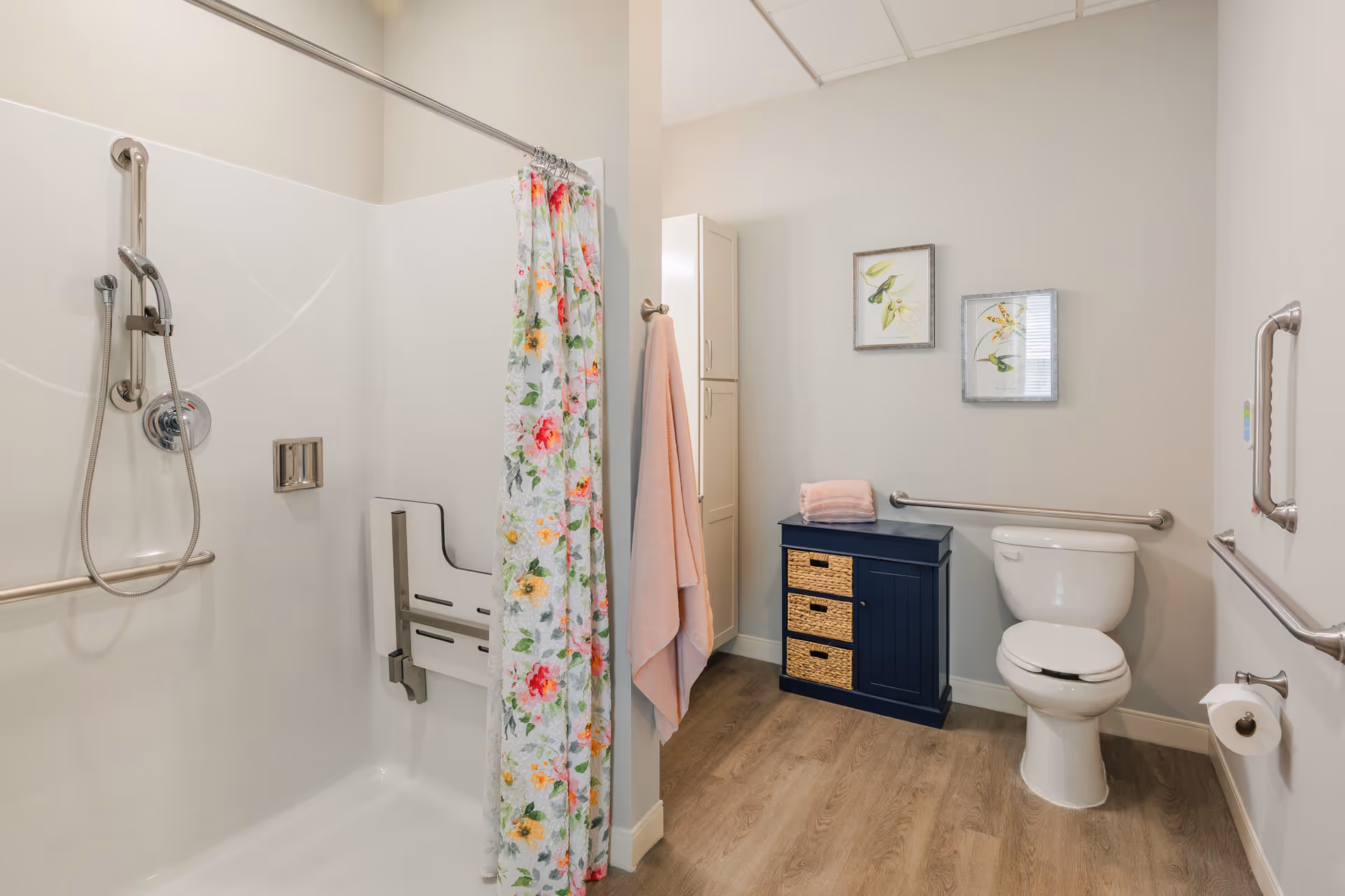 Bright accessible bathroom featuring a roll-in shower with a fold-down seat and floral curtain, a toilet with grab bars, and a navy storage cabinet.