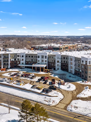 Aerial view of The Meadows Senior Living facility during winter, showing a large multi-story building surrounded by snow-covered ground and a parking area with several vehicles. The background includes a commercial area and a clear blue sky.