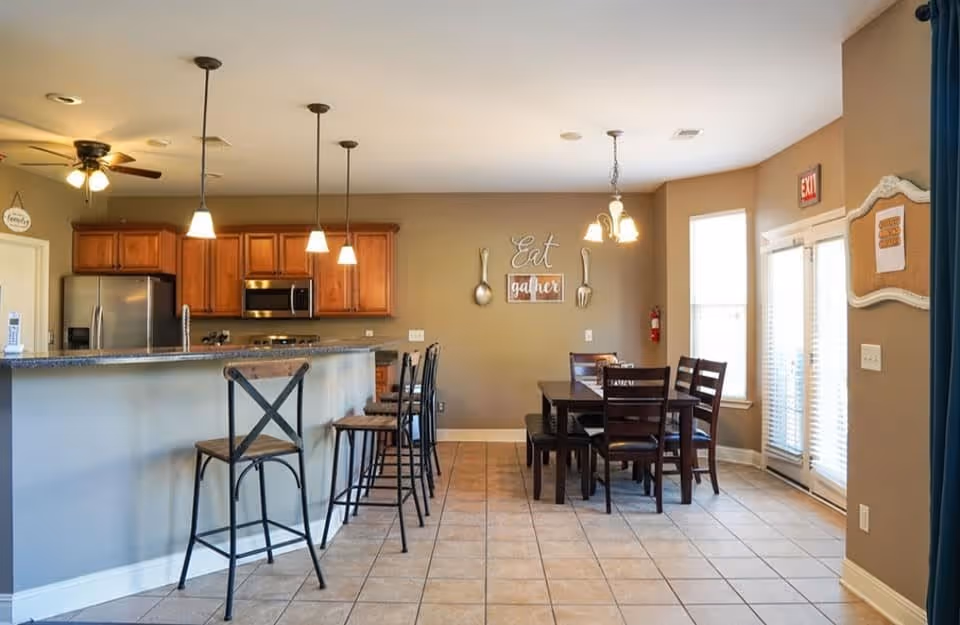 Open communal kitchen and dining area with a breakfast bar and stools on the left and a dining table and chairs on the right under pendant lights.
