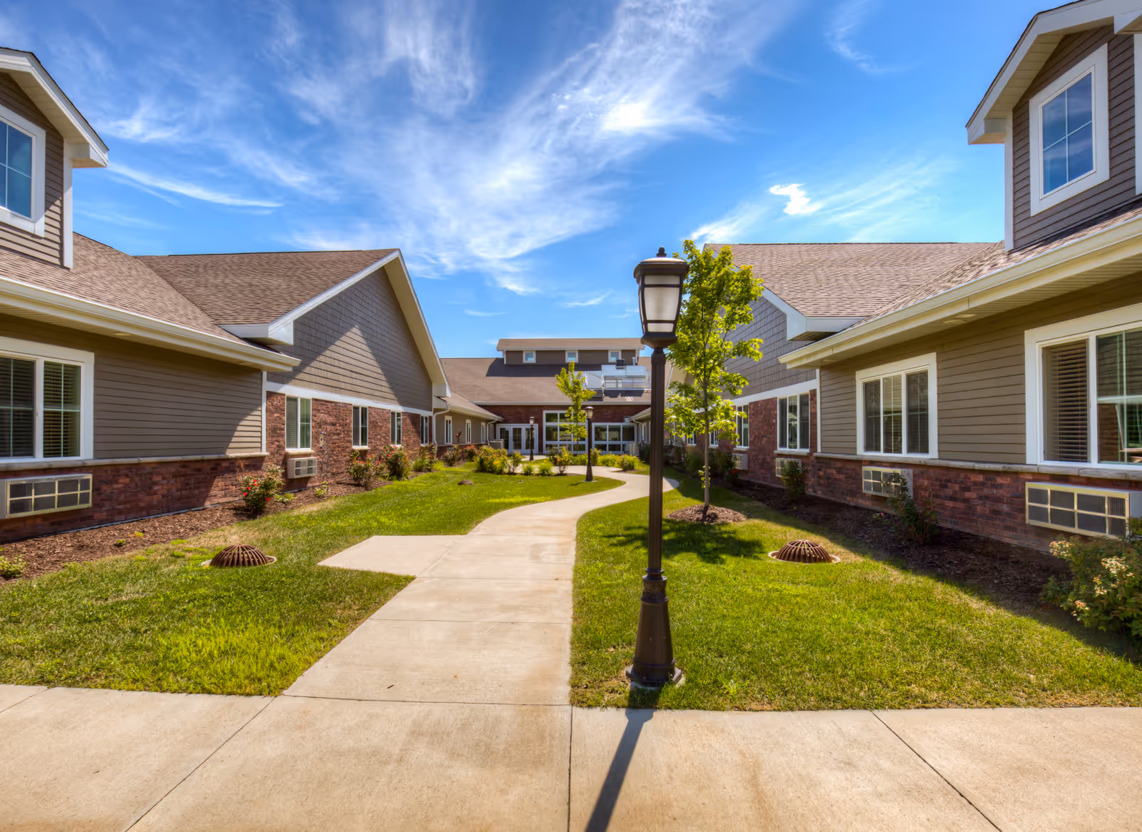 Sunlit courtyard with a paved walkway between two building wings and a central lamppost under a blue sky.