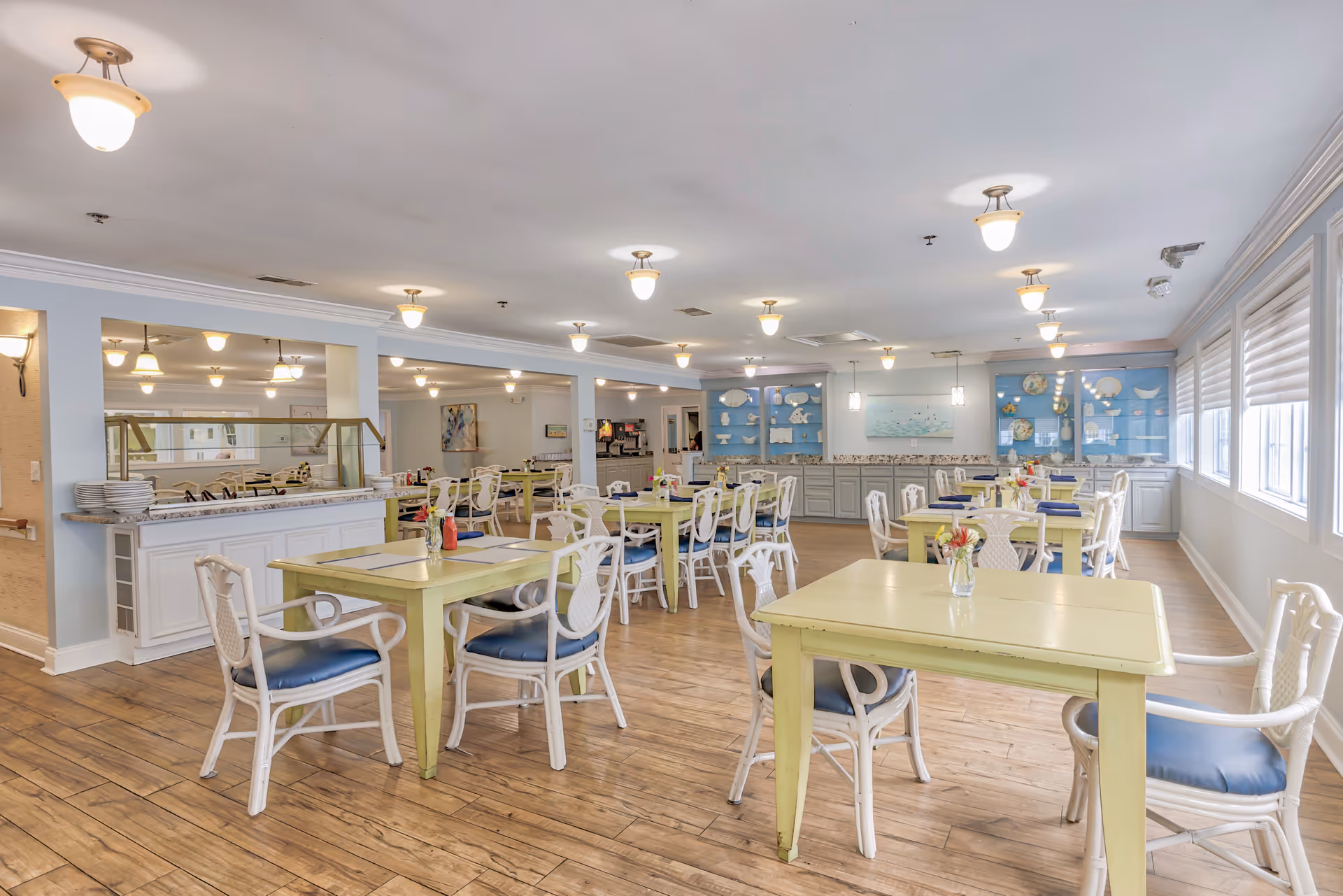 A bright and spacious dining room with multiple light green tables and white chairs with blue cushions. The room has wooden flooring, soft lighting from ceiling fixtures, and large windows with blinds on the right side. There are decorative shelves with plates and artwork on the far wall, and a counter area with plates stacked on the left side.