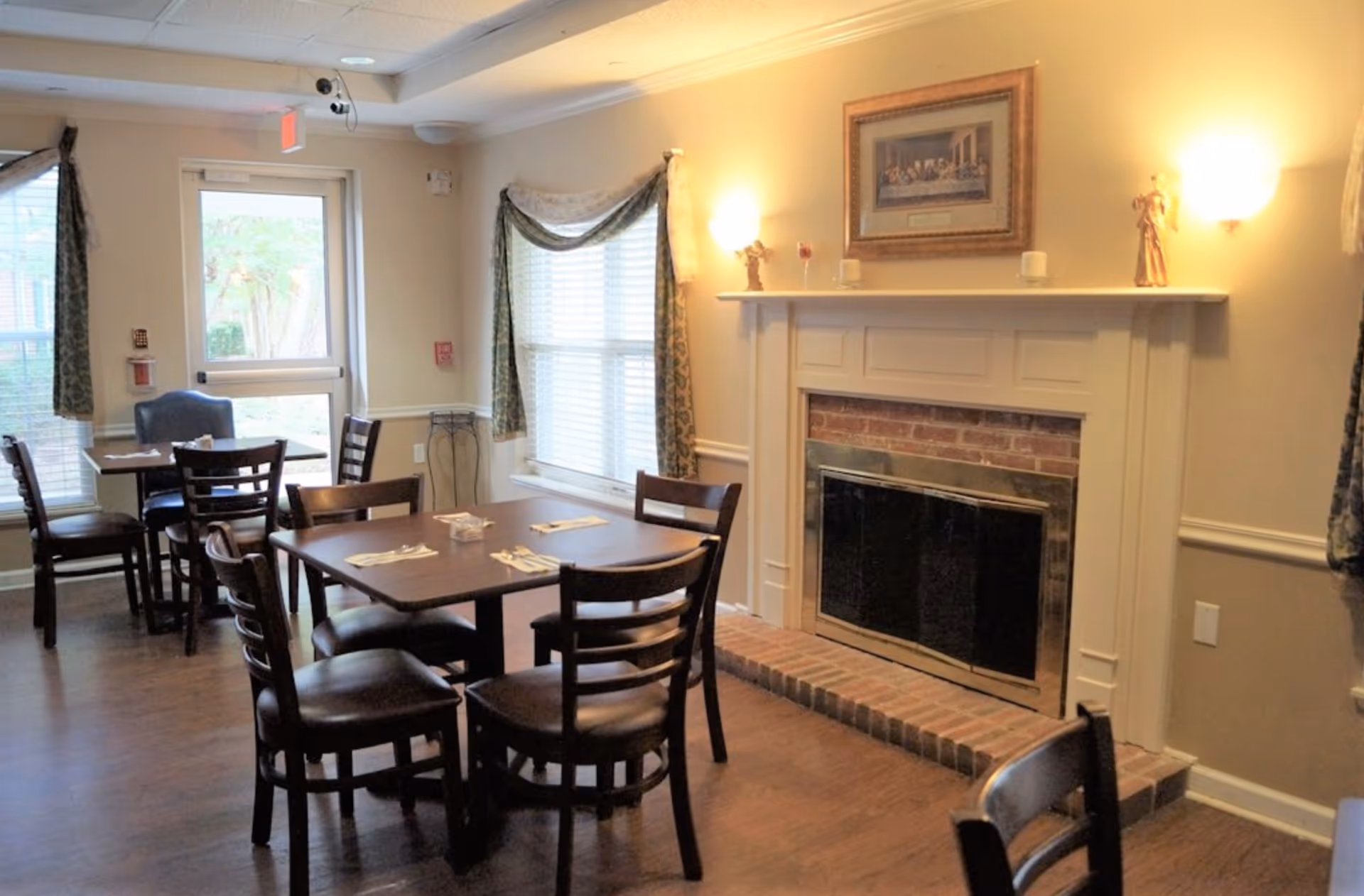 A cozy dining area with wooden tables and chairs, set with napkins and utensils. The room features a brick fireplace with a white mantel decorated with small figurines and candles. There are windows with patterned curtains allowing natural light to enter, and a door leading outside. The walls are painted beige, and the floor is wooden.
