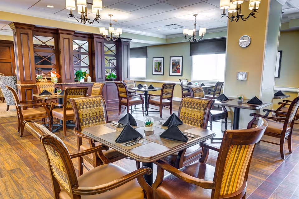 Well-appointed dining room with multiple wooden tables and upholstered chairs set with black napkins and chandeliers overhead.