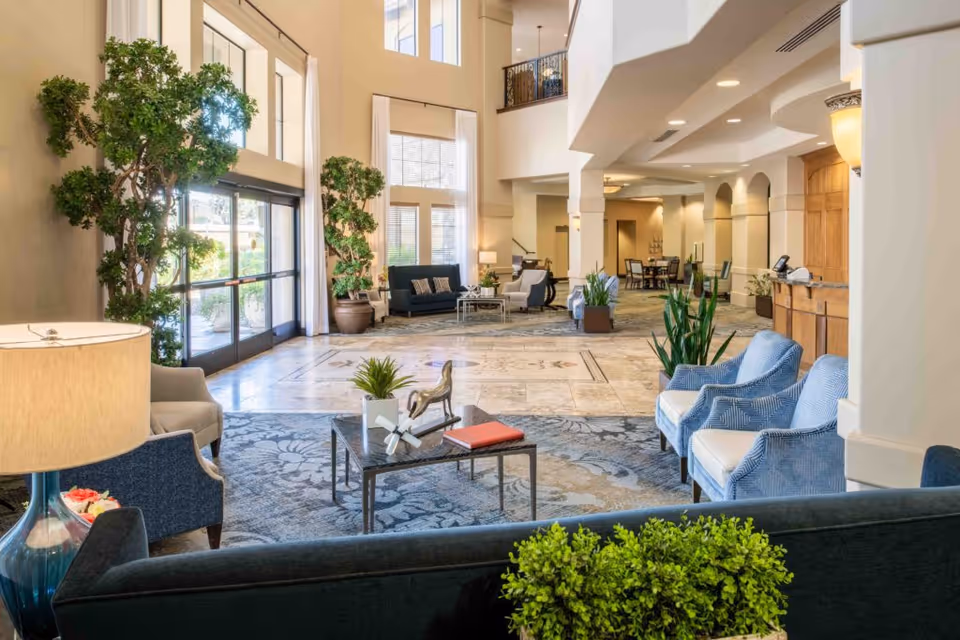 Bright and spacious senior living facility lobby with high ceilings, large windows, and glass doors letting in natural light. The room is furnished with comfortable seating including blue armchairs, a dark sofa, and a coffee table with decorative items. There are several large potted plants and a reception desk on the right side. The floor features a patterned carpet and tiled area, and there is a dining area visible in the background.