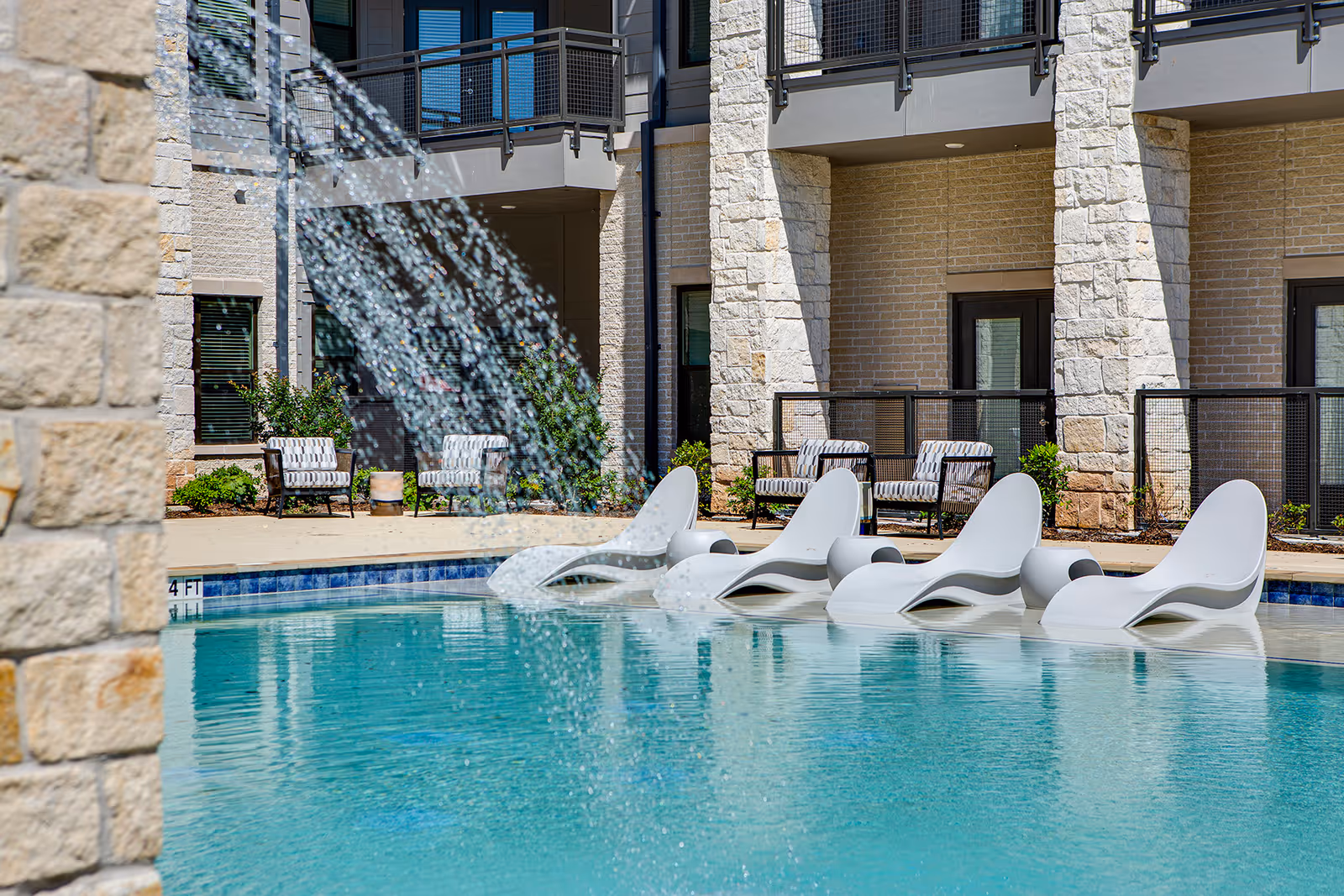 Outdoor swimming pool area with four white lounge chairs partially submerged in the water. Water is flowing from a stone structure into the pool. In the background, there are cushioned chairs and a building with stone and brick walls and balconies.