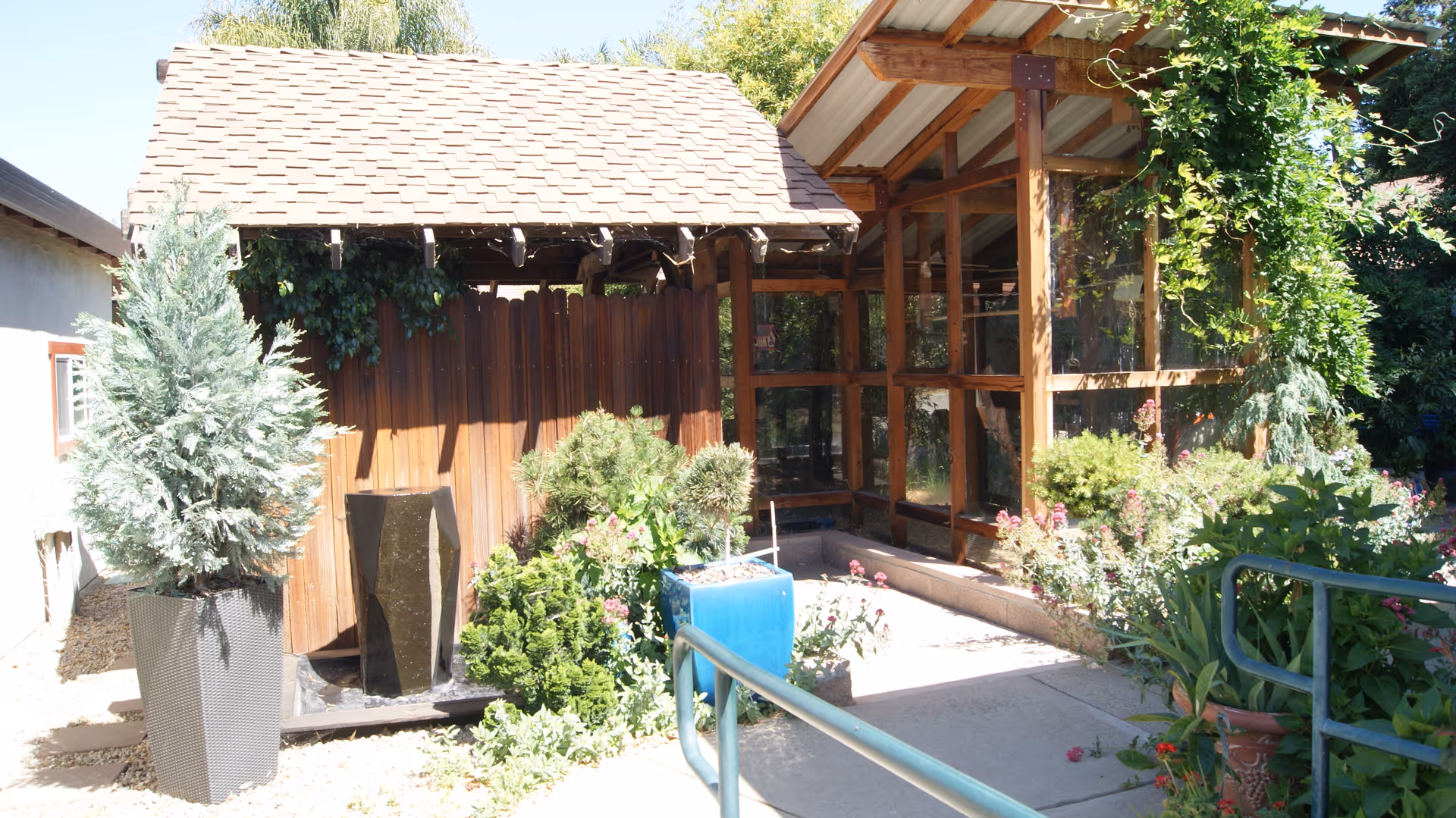 Sunny outdoor courtyard featuring a wooden pergola and glass-covered walkway surrounded by potted plants and shrubs.