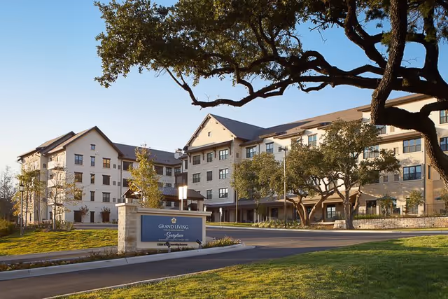 Exterior view of the Grand Living At Georgetown senior living facility building with multiple floors, surrounded by trees and greenery under a clear blue sky. A sign with the facility name is visible near the driveway entrance.