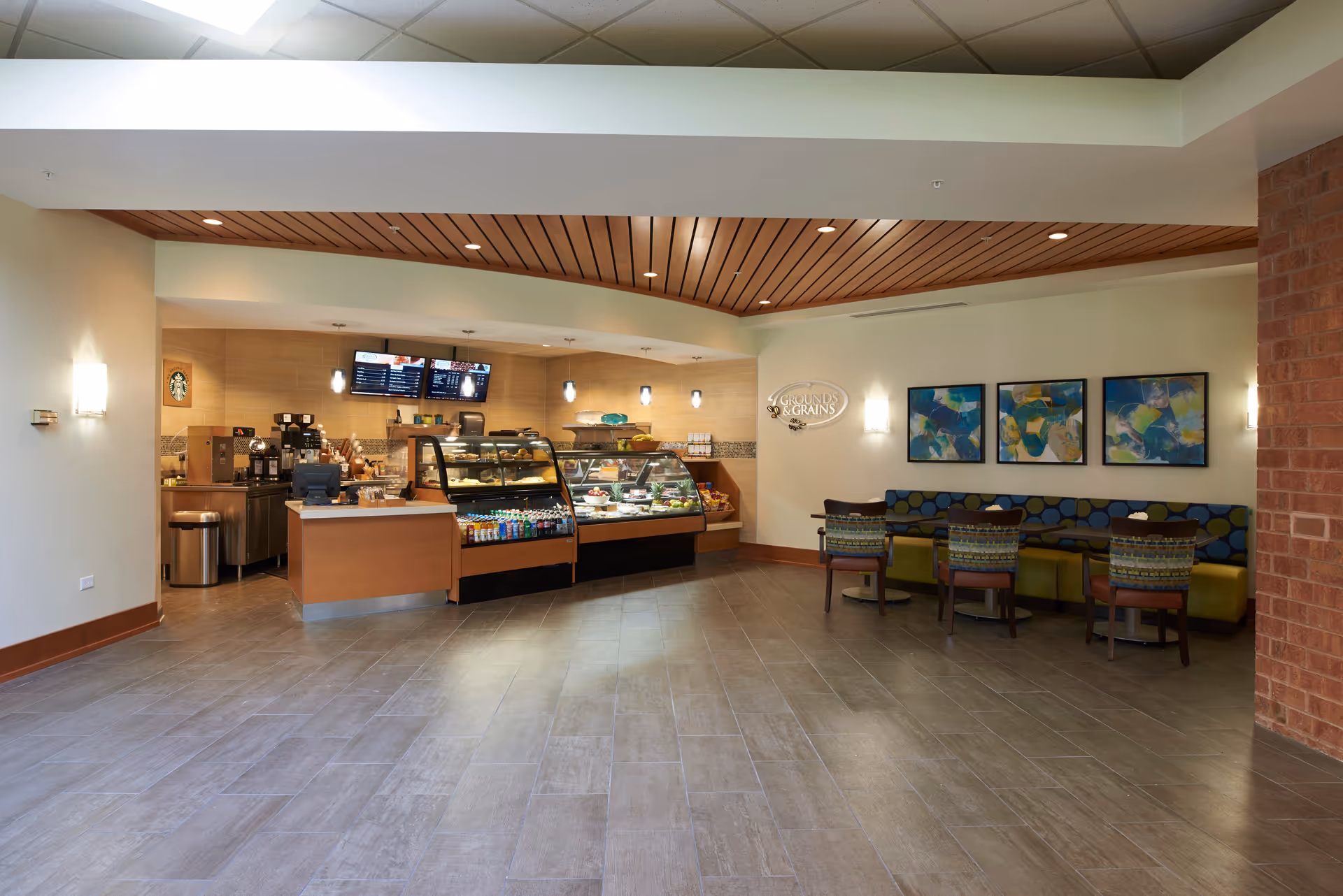 Interior view of a small cafe area inside a facility with a counter displaying baked goods and beverages. Behind the counter are coffee machines and menu screens. To the right, there is a seating area with a cushioned bench and chairs, along with three colorful abstract paintings on the wall. The ceiling has wooden paneling and recessed lighting.