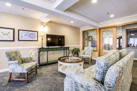 A cozy living room area with patterned armchairs and a sofa arranged around a round glass-top coffee table. A green console table with a flat-screen TV and a lamp is against the wall. The walls are light-colored with framed artwork, and there is a windowed door leading to another room.