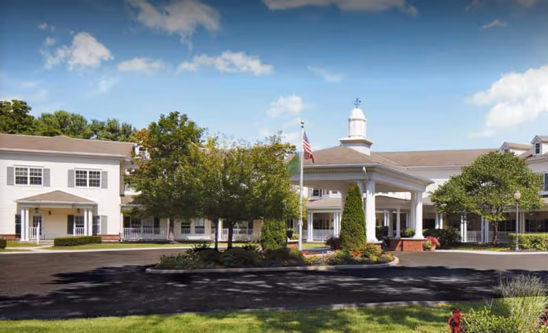 Exterior view of The Cottage at Litchfield Hills senior living facility showing a large white building with multiple windows, a covered entrance with columns, an American flag, trees, and landscaped greenery under a partly cloudy blue sky.