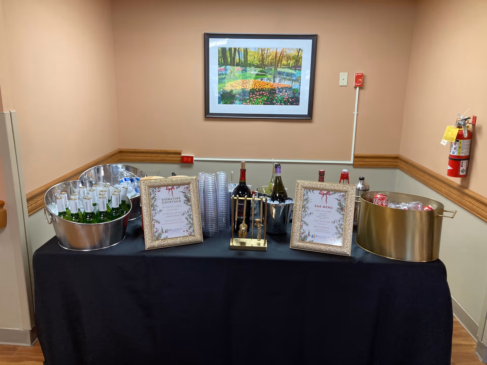 A black-covered refreshment table in a hallway with bottles, ice buckets, framed menus and plastic cups under a framed landscape picture.