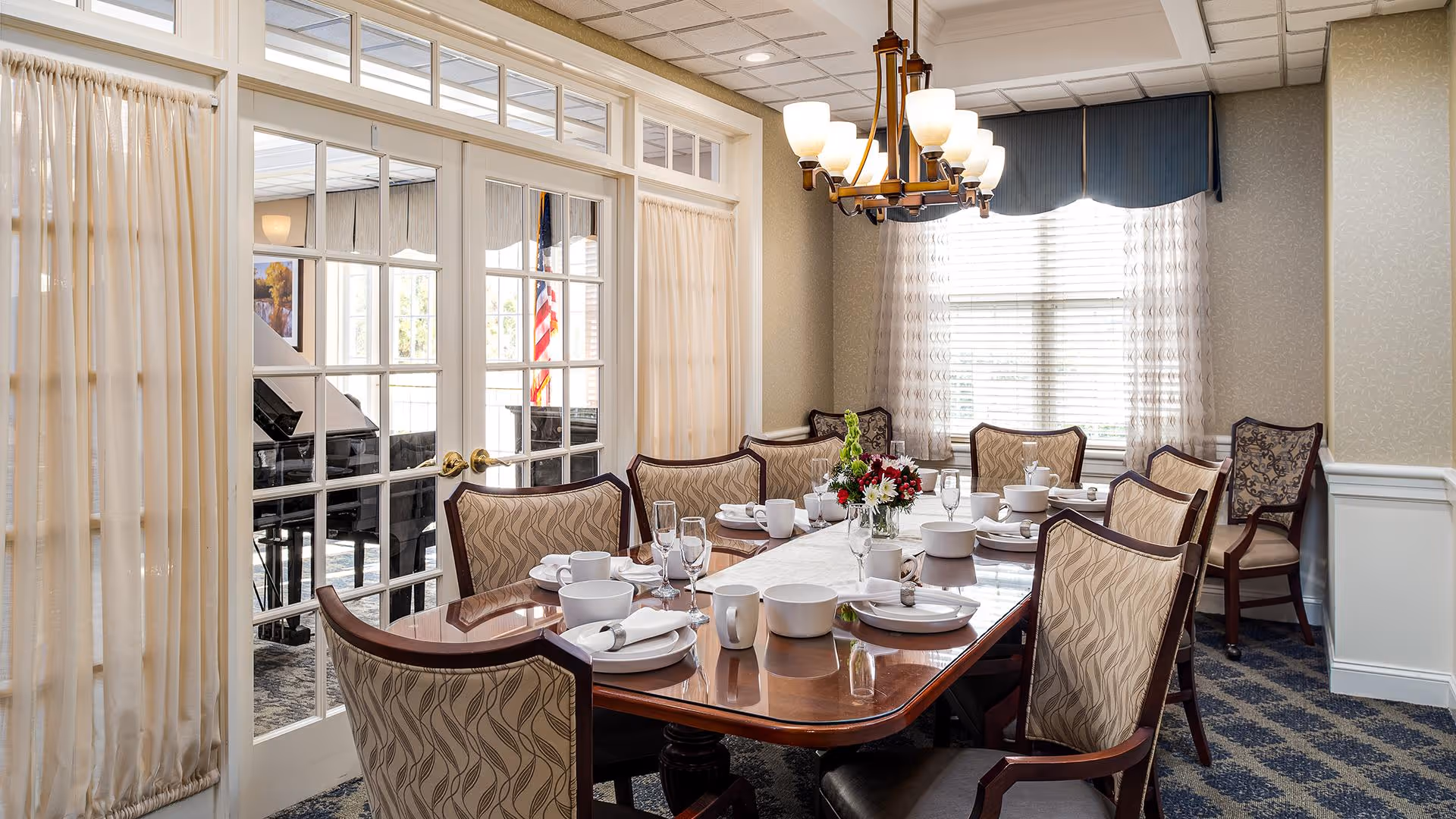 Formal dining room with a long set table, upholstered chairs, chandelier, and glass French doors revealing a piano in the adjacent room.