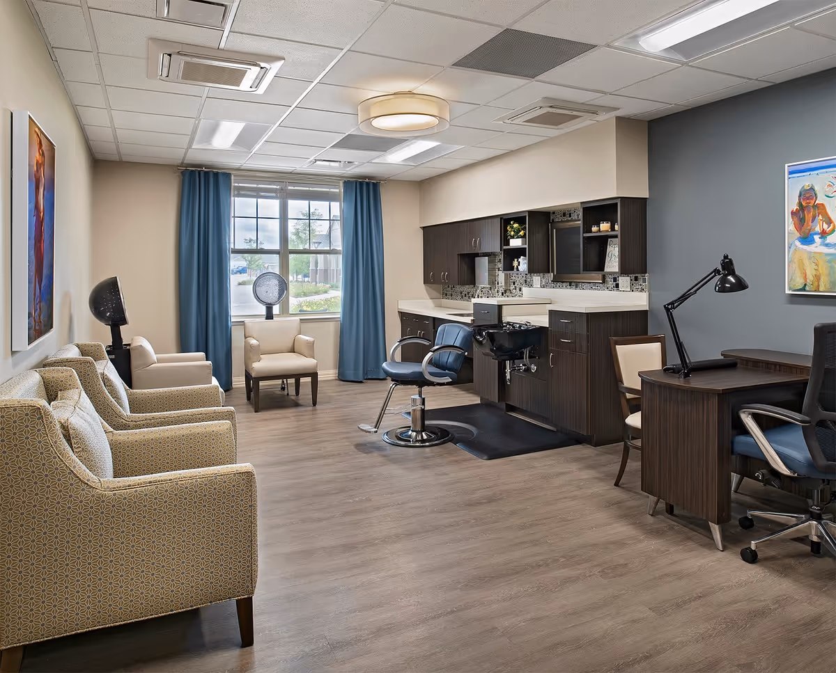 A well-lit salon area in a senior living facility with a styling chair, hair washing station, and cabinetry along one wall. There are several upholstered chairs along the opposite wall and a desk with a lamp and office chair. A large window with blue curtains lets in natural light, and colorful artwork hangs on the walls.
