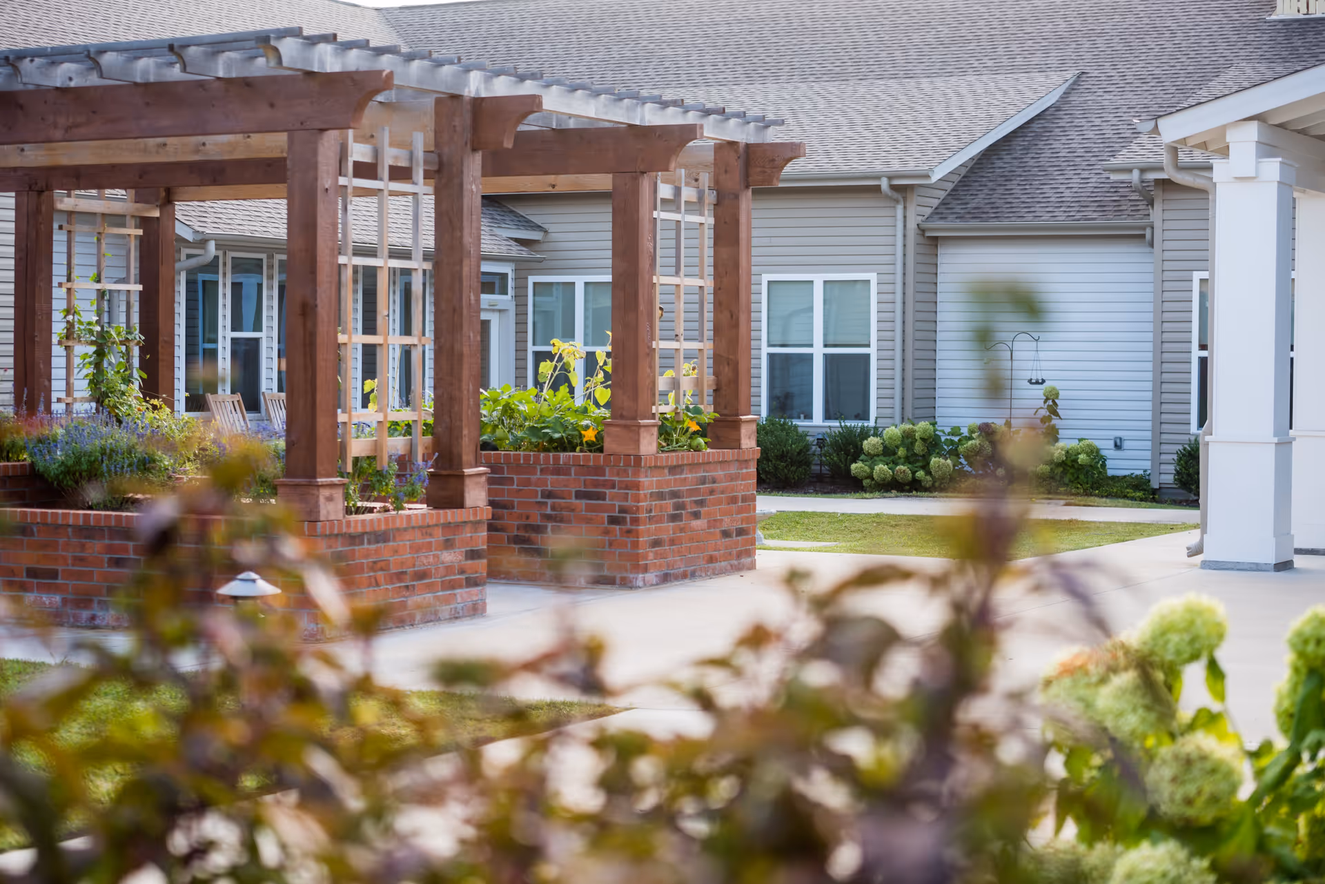 Outdoor garden area at a senior living facility featuring wooden pergolas with climbing plants, brick planters, and a paved walkway. The background shows the exterior of the facility building with windows and beige siding.