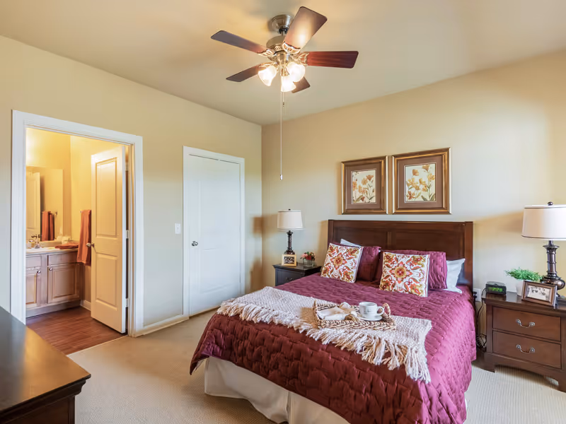 A cozy bedroom with a large bed covered in a burgundy quilt and decorative pillows. There are two nightstands with lamps on either side of the bed. Above the bed are two framed floral artworks. A ceiling fan with lights is mounted on the ceiling. An open door reveals a bathroom with a sink and towels.