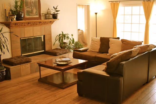 A cozy living room with a brown leather sectional sofa adorned with beige and brown cushions, a wooden coffee table with a decorative plate and books, a fireplace with a wooden mantel decorated with plants and framed artwork, and a large window with yellow curtains letting in natural light.