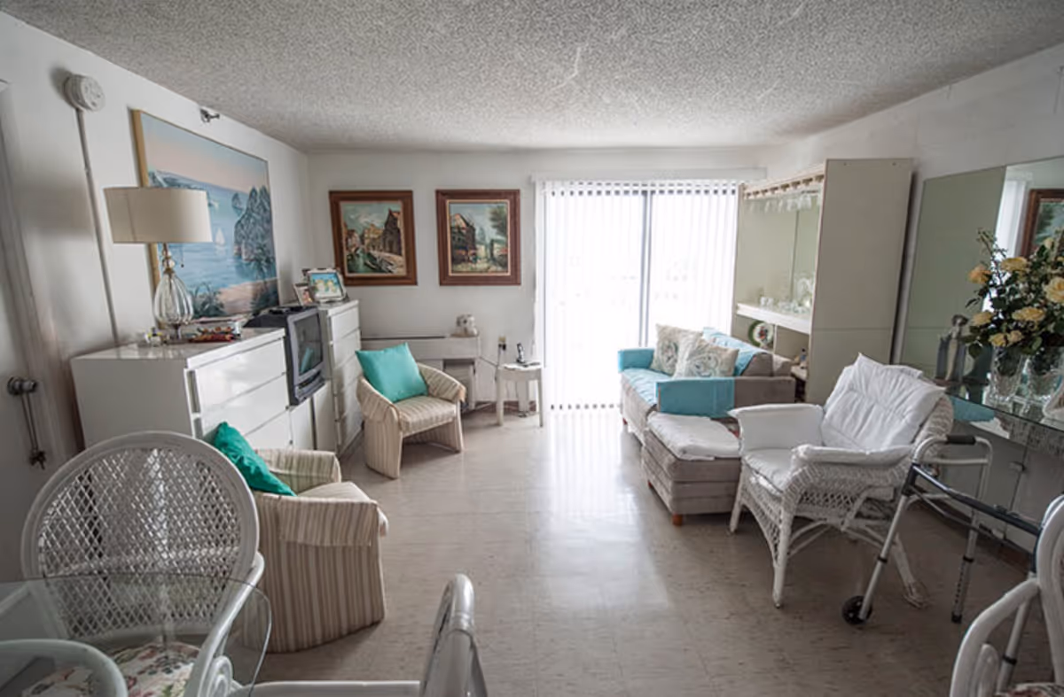 A bright living room with a glass dining table and wicker chairs in the foreground, cushioned armchairs with teal pillows, a small sofa with decorative pillows, a TV on a white dresser, framed paintings on the wall, a floor lamp, and a walker near a white wicker chair. Vertical blinds cover a sliding glass door letting in natural light.