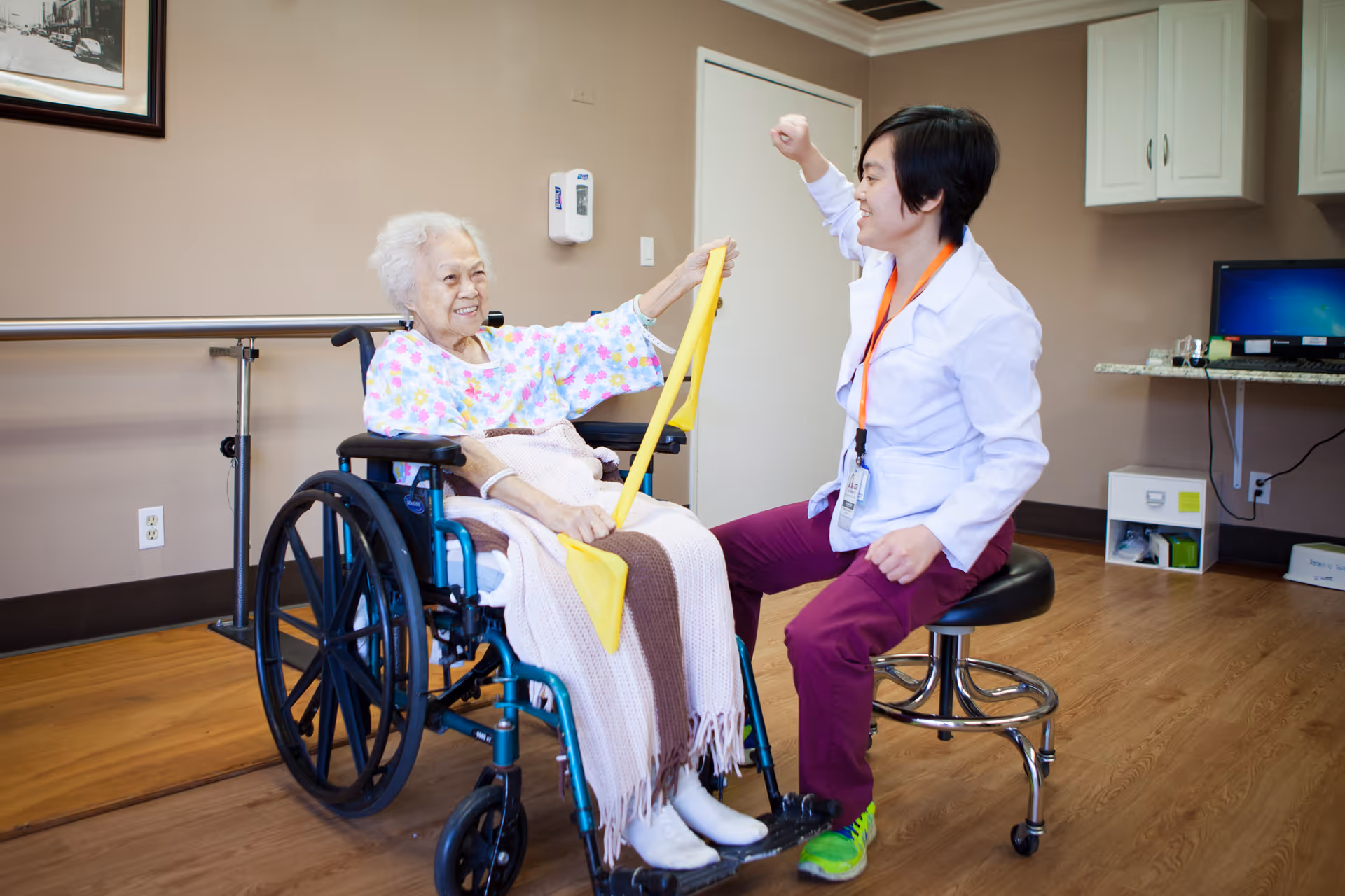 An elderly woman in a wheelchair is exercising with a yellow resistance band held by a healthcare professional in a white coat and purple pants, who is sitting on a stool and encouraging her. The room has wooden floors, beige walls, a computer on a desk, and medical equipment on the wall.