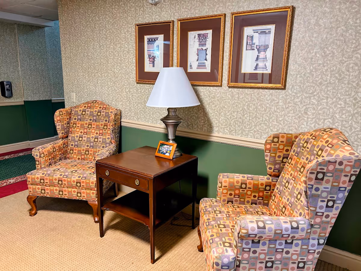 Two patterned wingback chairs flank a wooden side table topped with a lamp and small framed photo against a wallpapered wall with three framed prints.