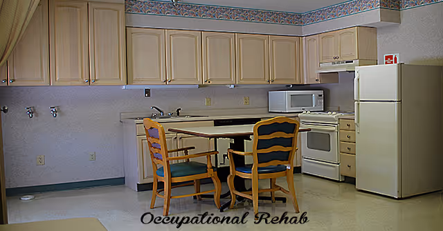 A kitchen area with light wood cabinets, a white refrigerator, a white stove with an overhead vent, and a microwave on the counter. There is a small table with two wooden chairs with cushions in front of the counter. The floor is light-colored, and there is a decorative border near the ceiling.