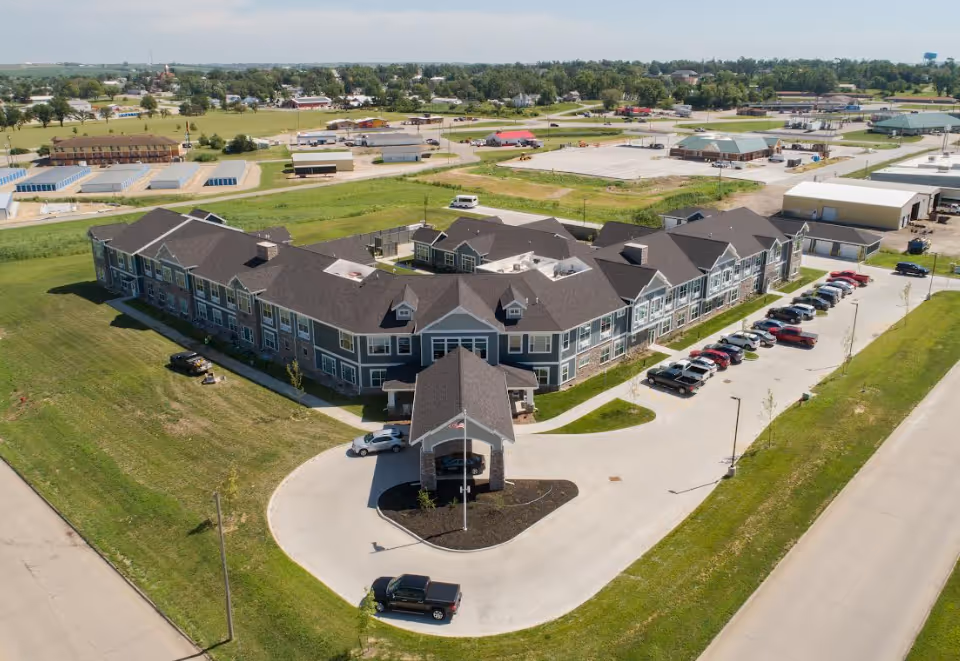 Aerial view of Birkwood Village Tama-Toledo, a large two-story senior living facility with a U-shaped building surrounded by green lawns and parking spaces filled with cars. The building has a covered entrance with a flagpole in front. The surrounding area includes roads, other buildings, and open spaces under a clear sky.