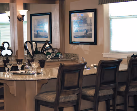 Interior view of a dining area with a granite countertop bar and four wooden chairs with cushioned seats. The countertop has several glasses and decorative items on it. The background shows two framed pictures on the wall and a window with blinds partially open.