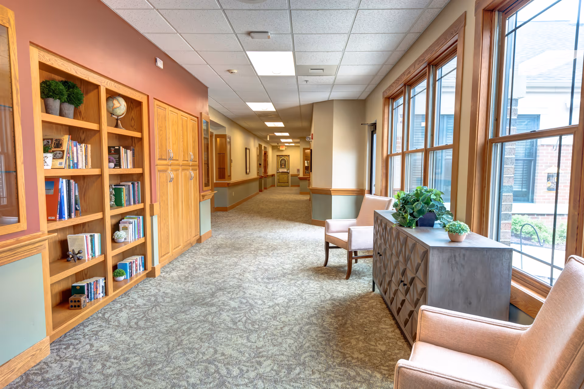 A bright hallway in a senior living facility with large windows on the right side letting in natural light. There are two upholstered chairs and a decorative cabinet with plants on top near the windows. On the left side, there is a built-in wooden bookshelf filled with books and decorative items. The hallway has carpeted flooring and a ceiling with recessed lighting.
